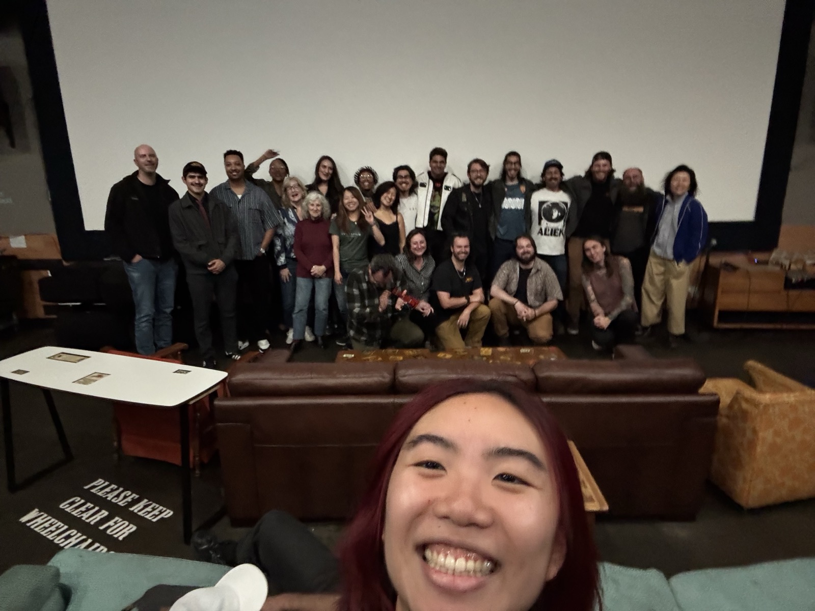 A wide selfie taken from the front row with the whole cast and crew grouped in front of the screen behind